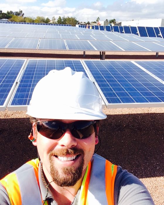 Selfie in a white hard hat and safety glasses in front of rows of fixed-tilt panels, palm trees behind.