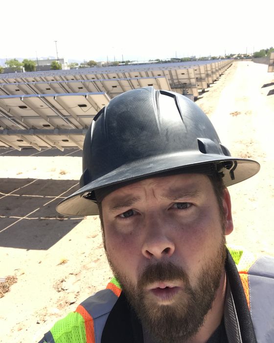 Close selfie in a black hard hat, looking into the sun, with the backs of panel rows behind.