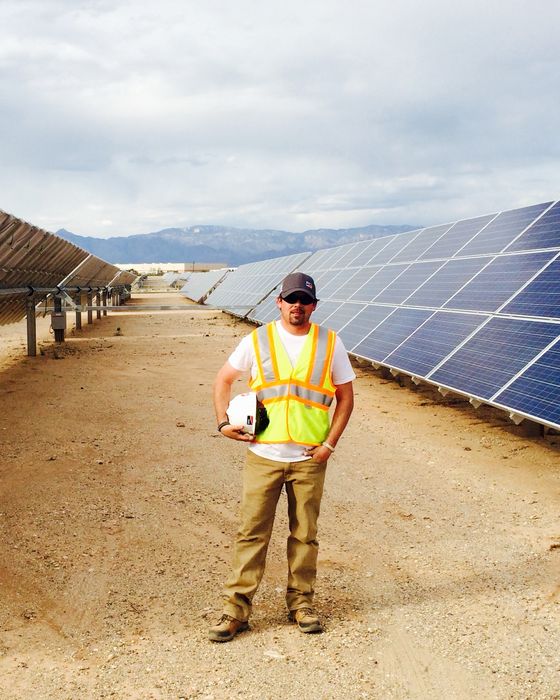 Standing in the aisle between two tracker rows, holding a hard hat, mountains in the distance.
