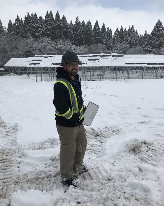 Standing in snow with a tablet, snow-covered array and cedar trees behind.