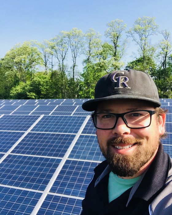 Selfie in a ball cap and glasses in front of a large array, trees in spring leaf behind.