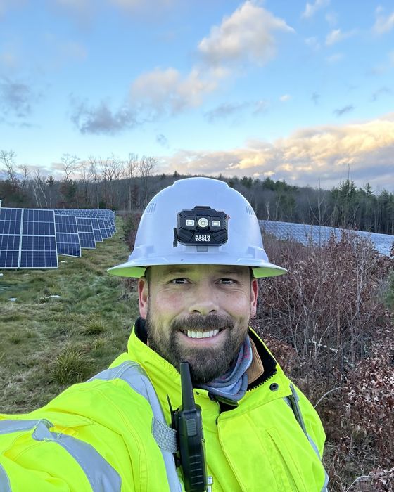 Selfie in a white hard hat with a headlamp, array rows and late-autumn woods behind.