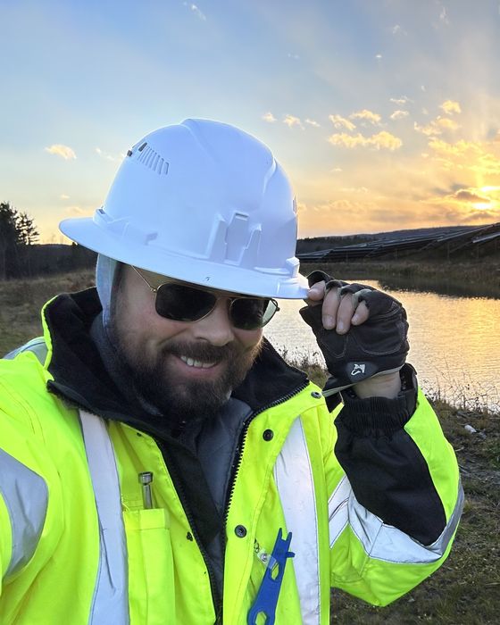 Tipping a white hard hat by a pond, a hillside array and sunset behind.