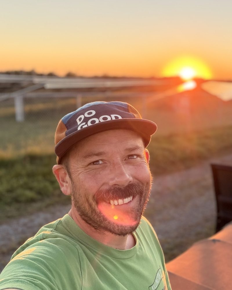 Casey Schuyler smiling in a ball cap, with a solar array and the setting sun behind him.